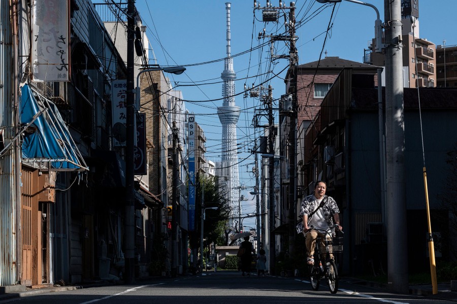 A man rides a bicycle past older buildings in Tokyo, with a tall modern skyscraper in the background.