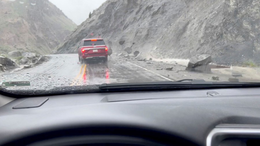 A view from inside a car, looking at another car ahead of it that is being battered by several small boulders that fell from a steep hill.