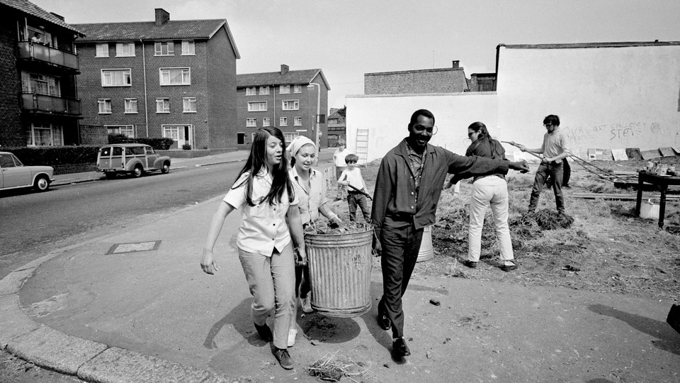 Image of two young women and one young man carrying a trash can together down a neighborhood sidewalk.
