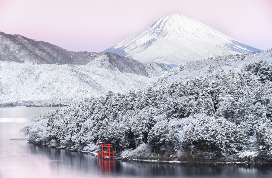 A red wooden gate stands along a lakeshore, with a snow-covered forest and mountains in the background.