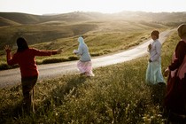 Girls walking along a road in Palestine, surrounded by rolling green hills, the sun beginning to set