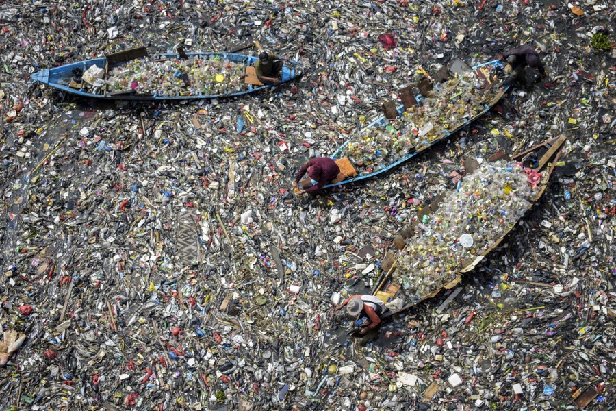 An aerial view of people in three small boats, all full of plastic items, floating in a garbage-choked river.
