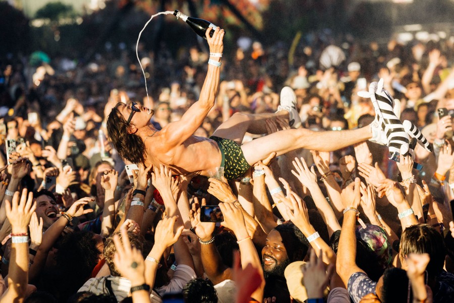A performer lies on his back, being held up by the many hands of a concert audience, while trying to drink from a champagne bottle.