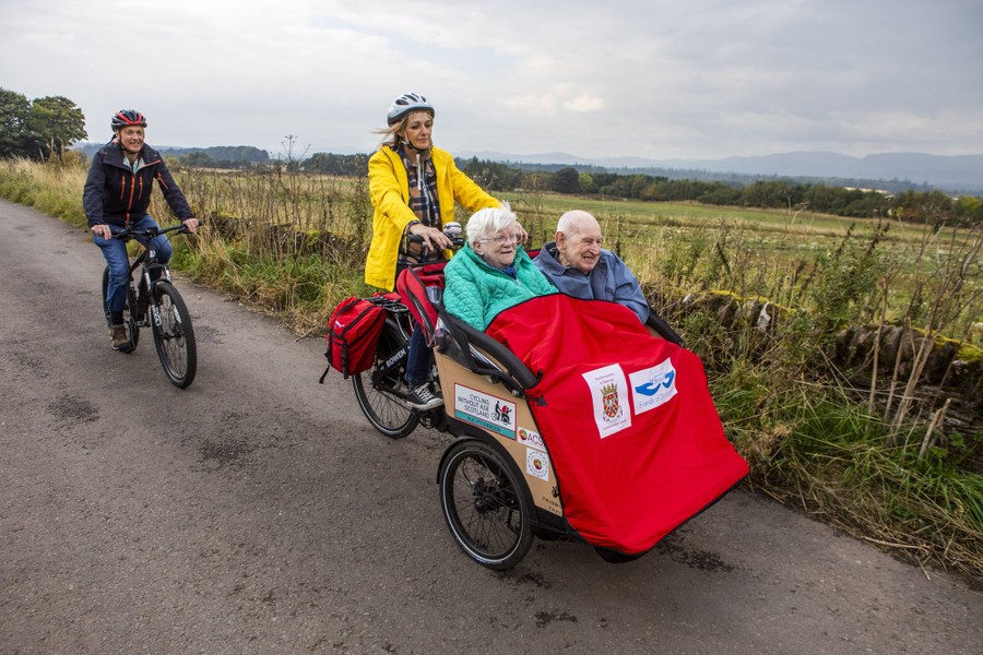 A person rides a bicycle on a path, following another person pedaling a three-wheel bike, carrying two older passengers in front.