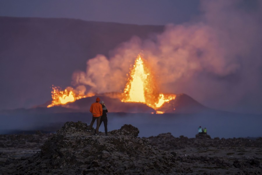 Several people stand and sit on a rocky plain, looking toward a distant fountain of lava erupting from a crater.