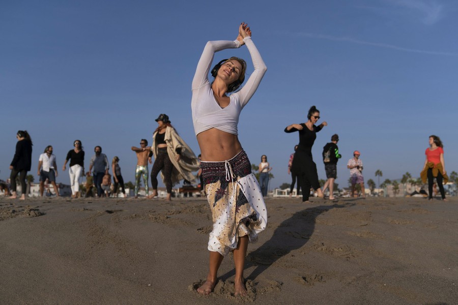 A woman wearing headphones dances on a beach; others dance nearby, spaced out from one another.