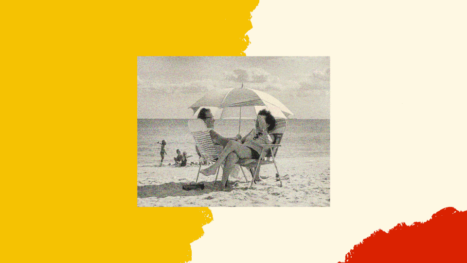 A black and white photo shows a white couple siting in folding chairs on the beach, facing one another and sharing the shade of a single umbrella.
