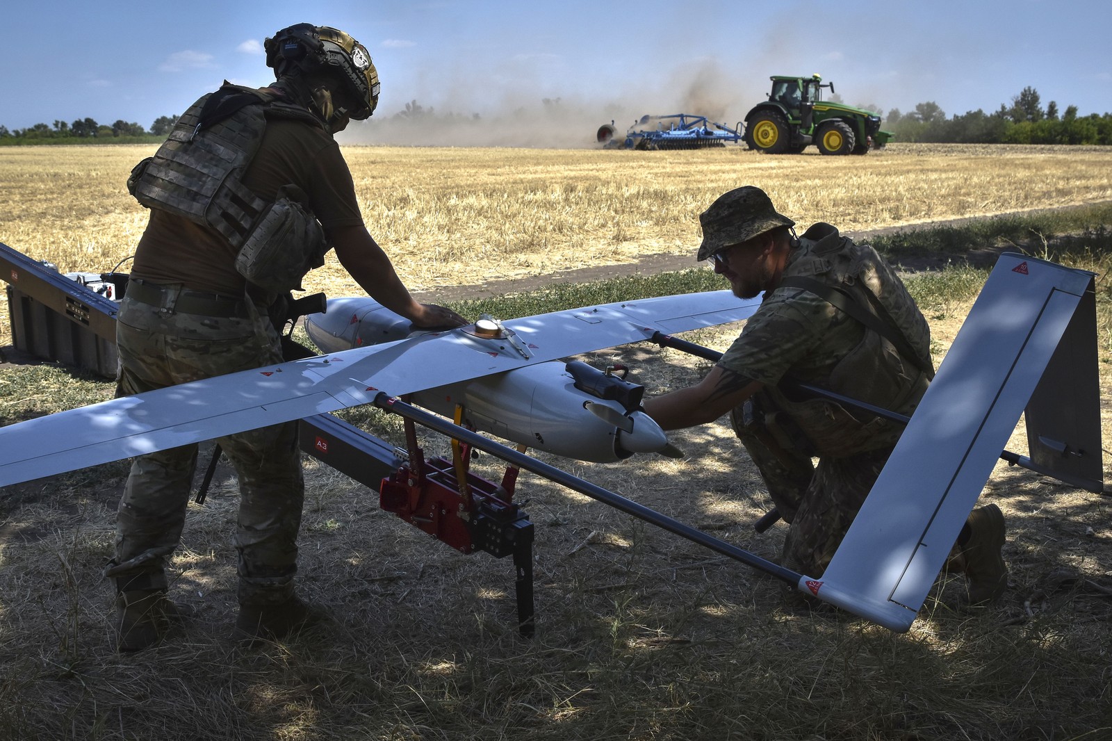 Two soldiers prepare a fixed-wing drone for a launch, beside a farm field with a tractor operating in the background.