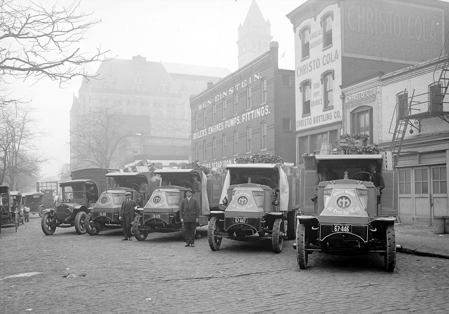 Two men stand beside a line of five 1920s-era delivery trucks parked in a street.