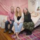 Two parents and their young adult daughter sit on a bed in a college apartment.