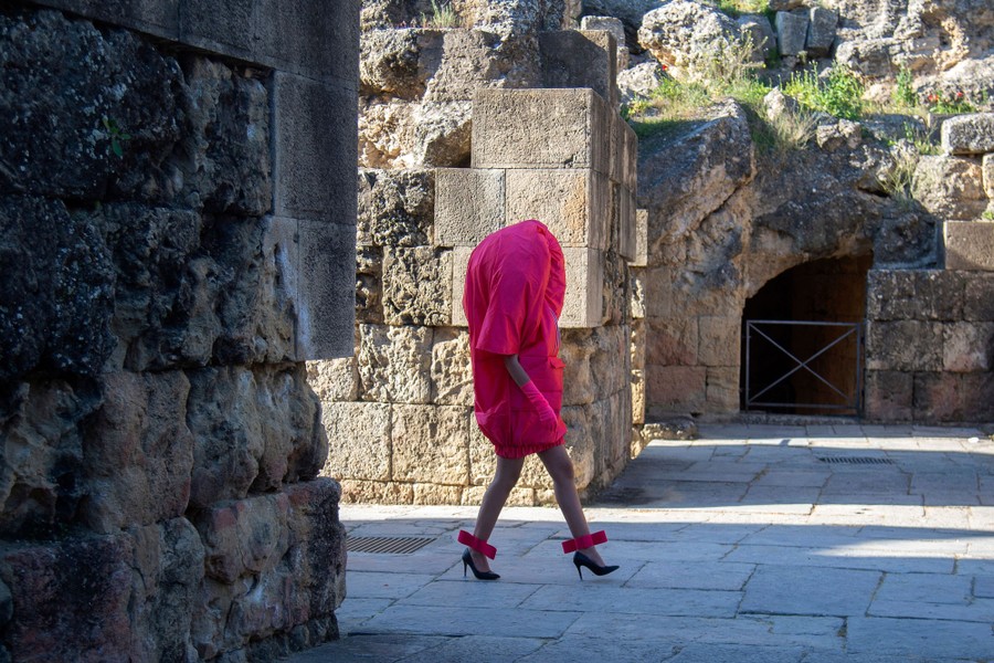 A model walks among ancient ruins, wearing a colorful oversize garment.