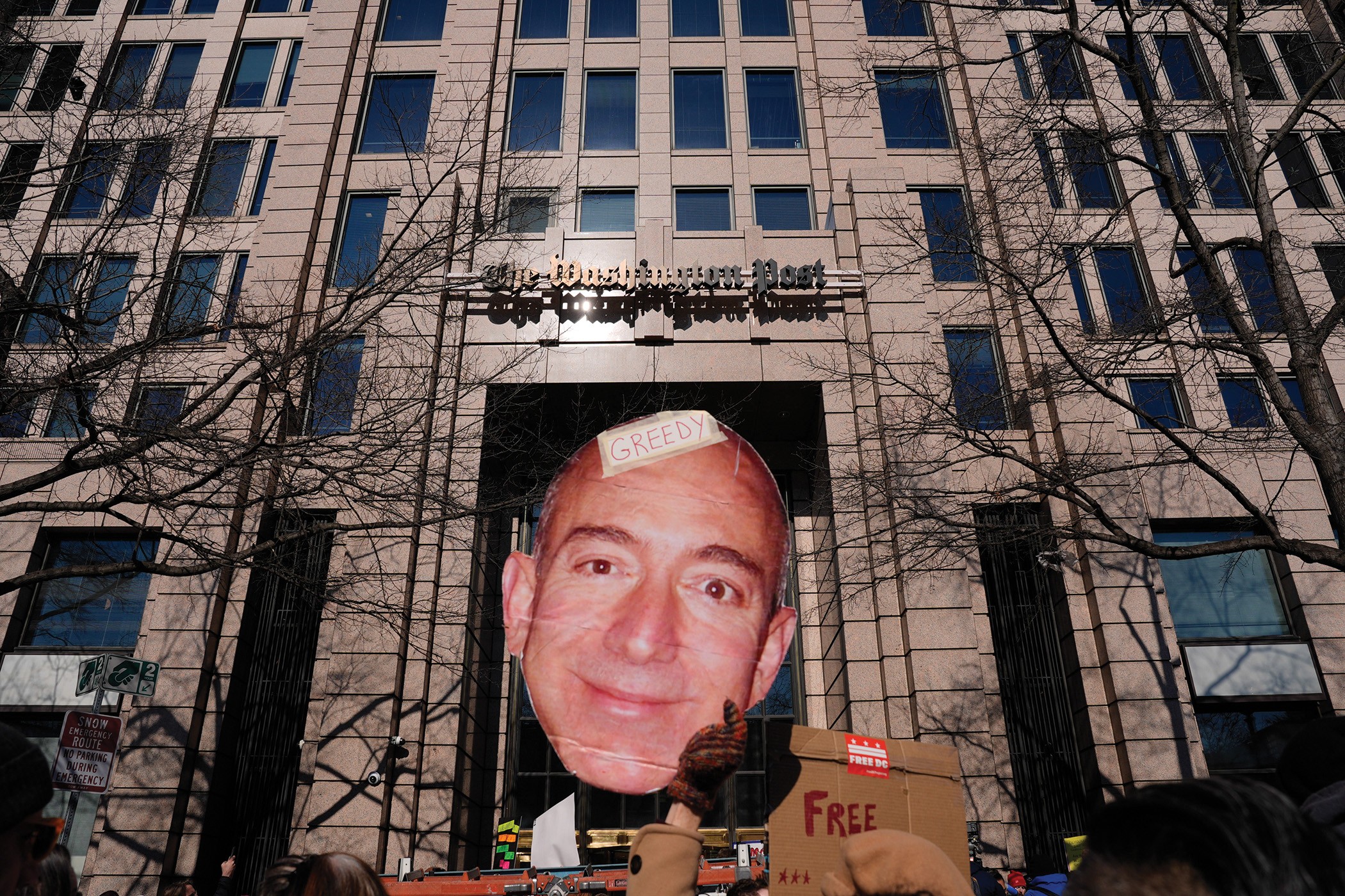 photo of rally with Washington Post building in background and large cardboard protest sign with Jeff Bezos's face on it