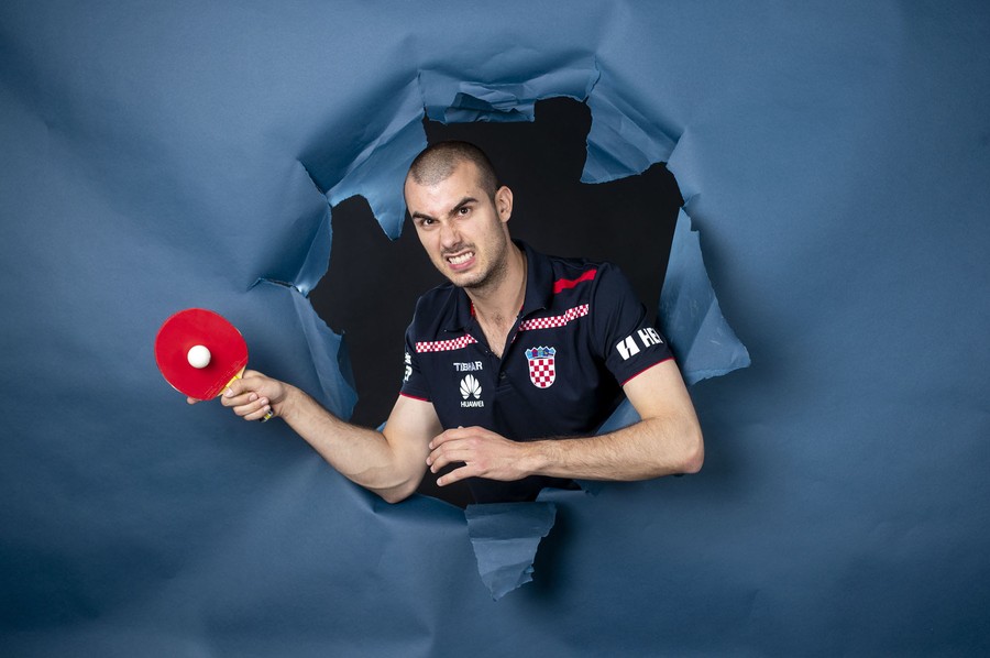 A table tennis player poses with a paddle and ball, appearing to burst through a hole in a large sheet of paper.