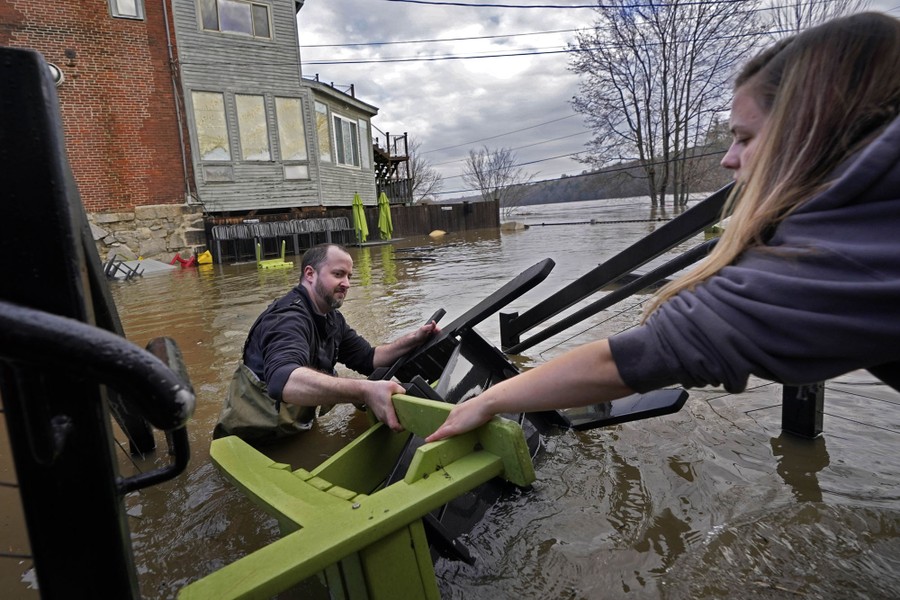 Two people work in hip-deep floodwater to recover wooden chairs from a patio.