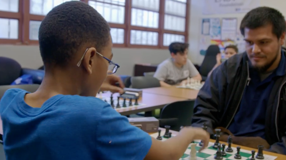 A young boy in a blue shirt and glasses plays chess with a slightly older boy
