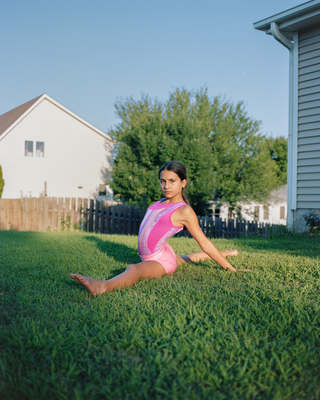 girl in ponytail and pink leotard does a split on green grass in a fenced yard next to house