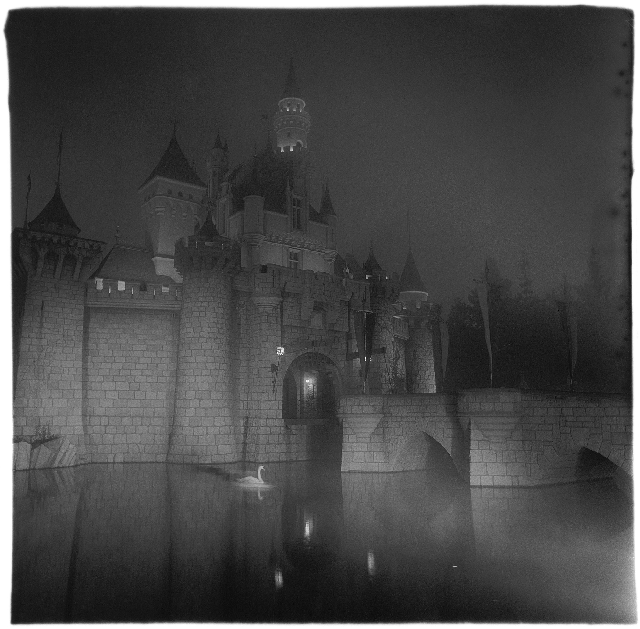black-and-white photo of lit tower and water-filled moat of Disneyland's Cinderella castle at night, with a swan swimming in the moat