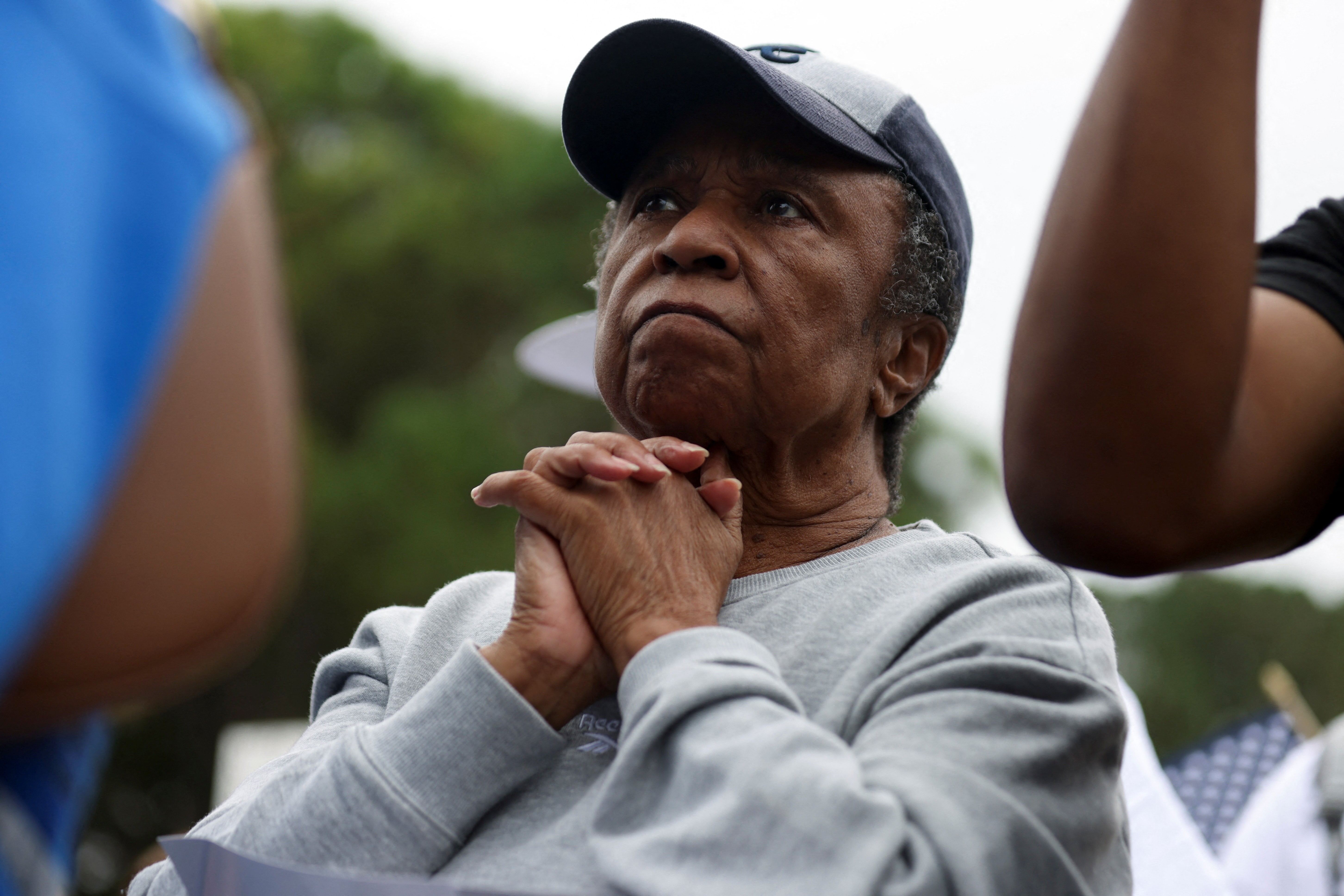 A person watches protesters march past, hands clasped beneath their chin.