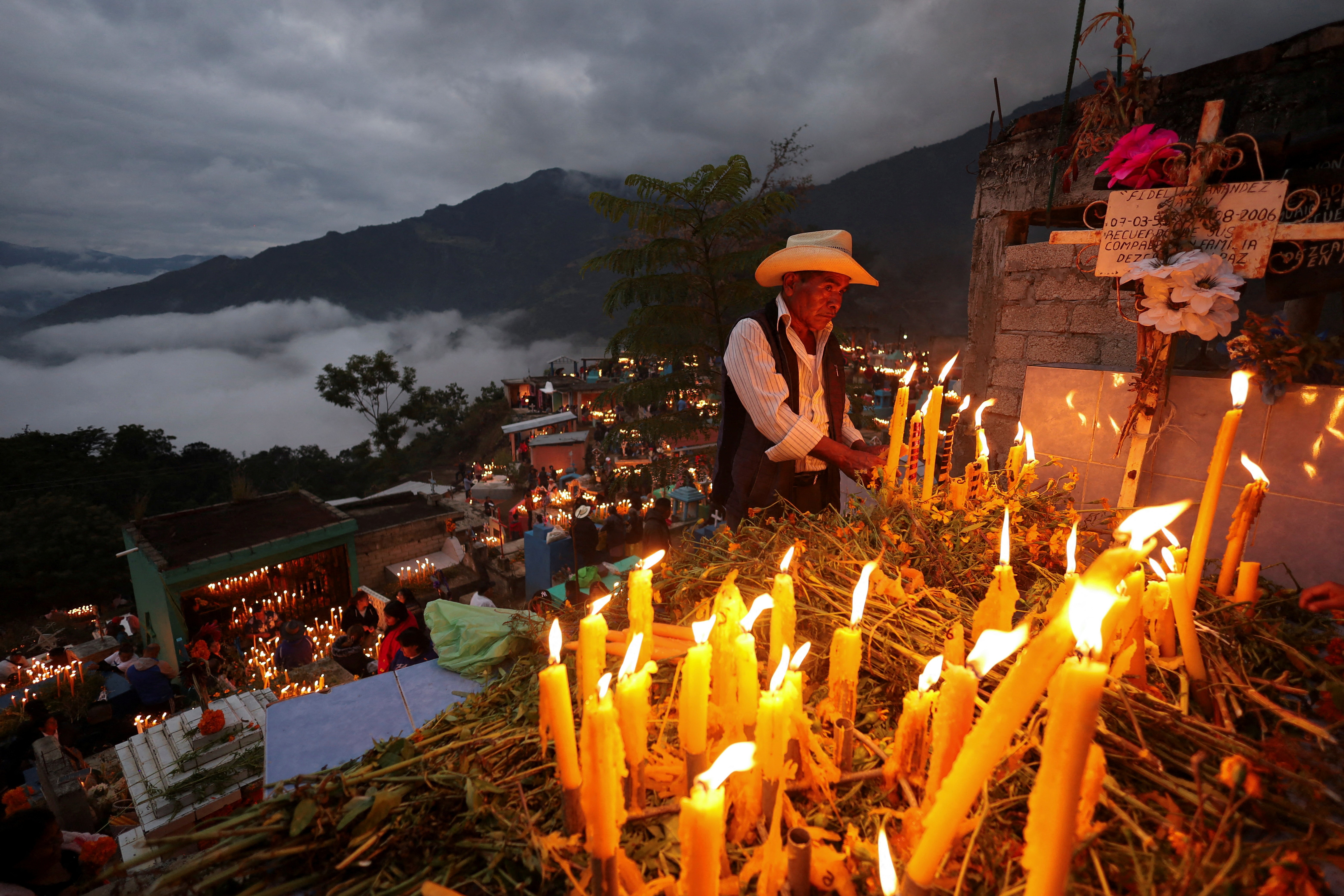 A man lights one of many candles burning on a grave, with many other candles on graves visible below on a hillside cemetery.