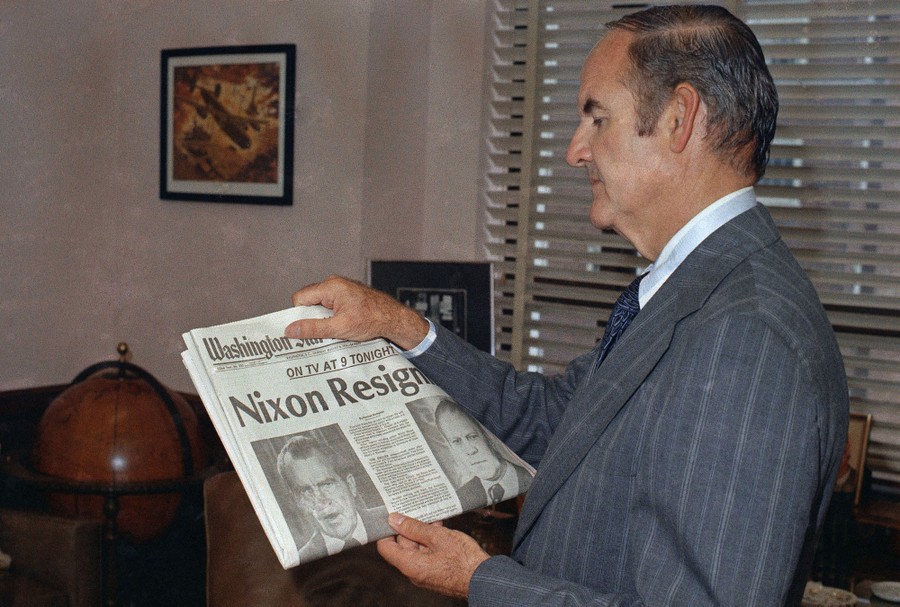 A person holds up a newspaper with "Nixon Resigns" as the headline.