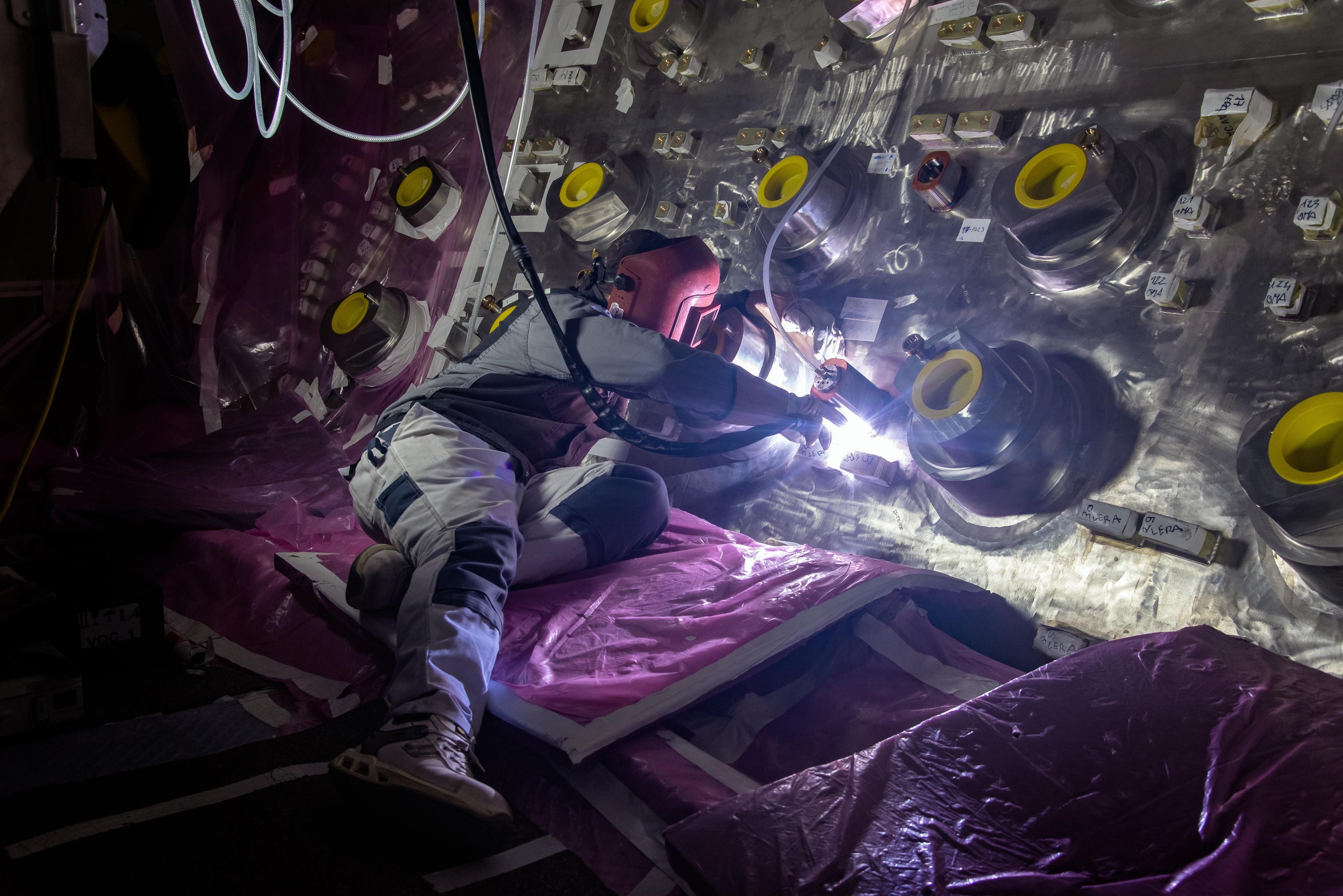 A worker welds an object onto the interior wall of a fusion reactor component.