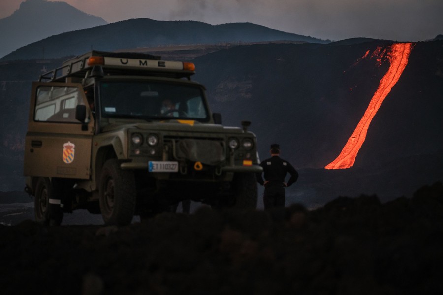 A ribbon of lava flows down a hillside in the distance, as people watch near a vehicle.
