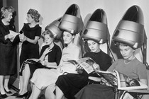 A group of women sit underneath hair dryers and read magazines at a hair salon.
