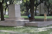 Two passersby take photos of a pedestal where a statue once stood. 