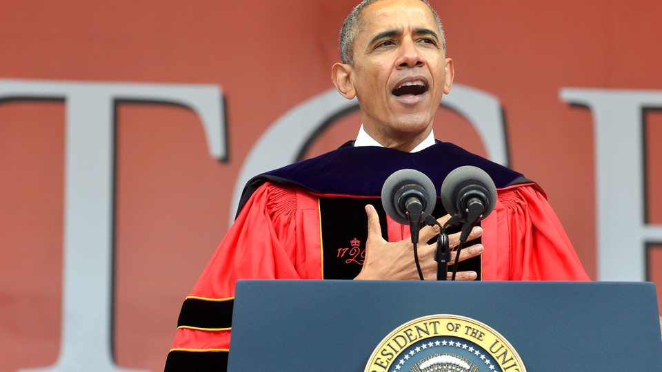 Barack Obama stands at a microphone wearing a graduation gown.