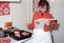 A woman reads a cookbook next to meat cooking on the stove.