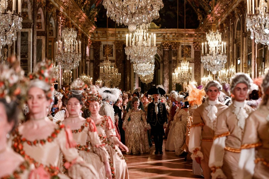 Dozens of people wearing baroque-style costumes attend a ball inside a very ornate palace ballroom.