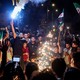 Syrians in a crowd light sparklers and wave flags on the street.