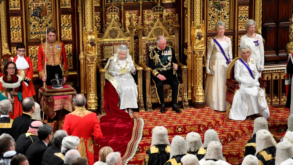 Queen Elizabeth and Prince Charles sit on golden thrones, surrounded by members of Parliament and other officials in both formal and official attire..