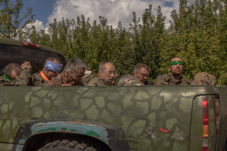 At least six blindfolded soldiers sit in the bed of a military pickup truck.