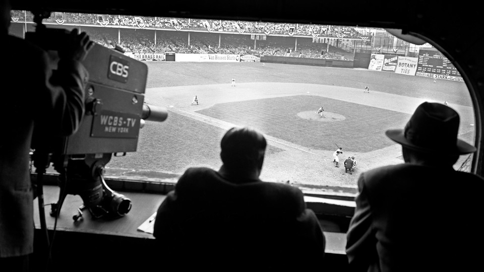 A black and white photograph of the CBS press box with one person monitoring a camera on the left, and two other figures, looking onto a baseball diamond