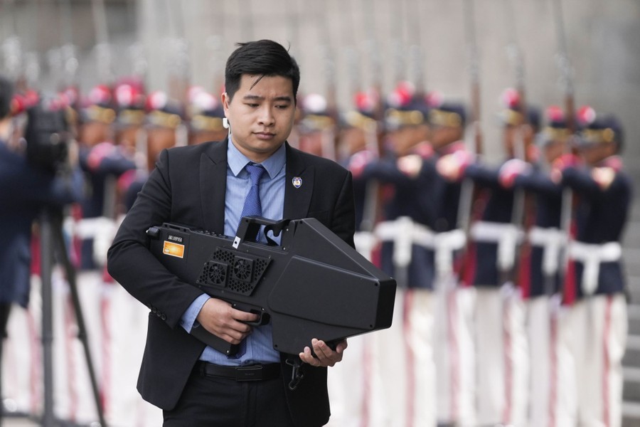 A member of a security detail carries a large black device, roughly shaped like a bulky rifle, near a line of soldiers in dress uniform.