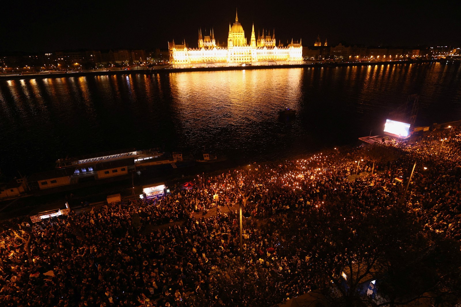 An aerial view of a crowd gathered on the Danube River, with Hungary's parliament building lit up on the opposite side of the river.