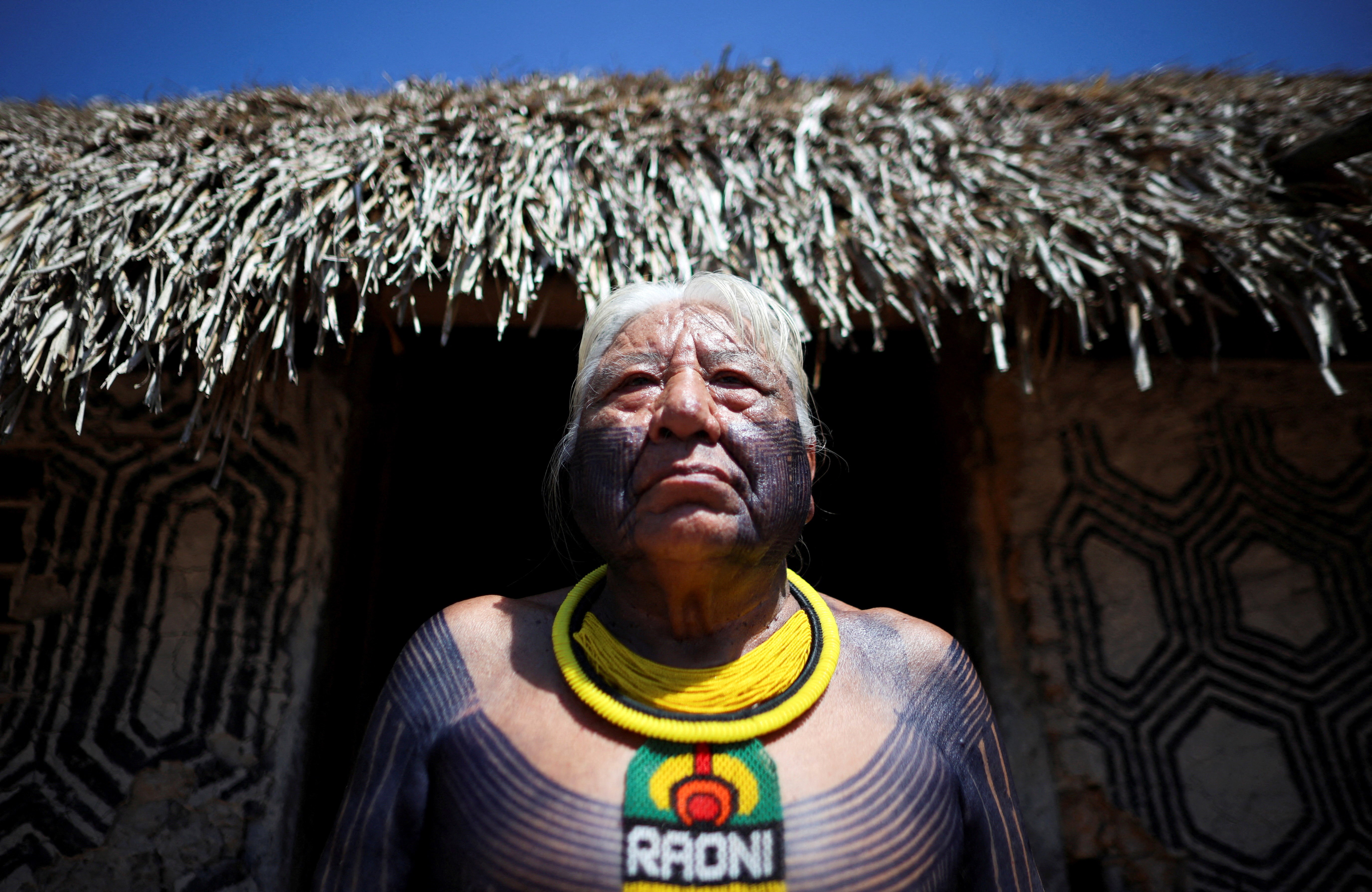 An indigenous man in Brazil poses in front of a building with a thatched roof. 