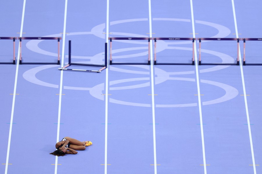 A runner lies on a track, hands to their face, with a toppled hurdle in the background.