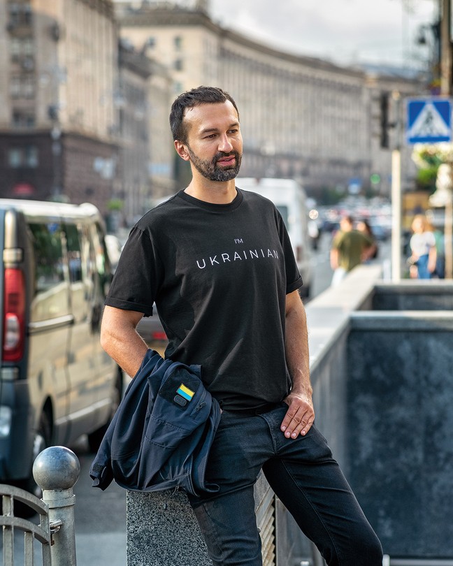 photo of man wearing "I'm Ukrainian" t-shirt, leaning against wall with busy city street in background