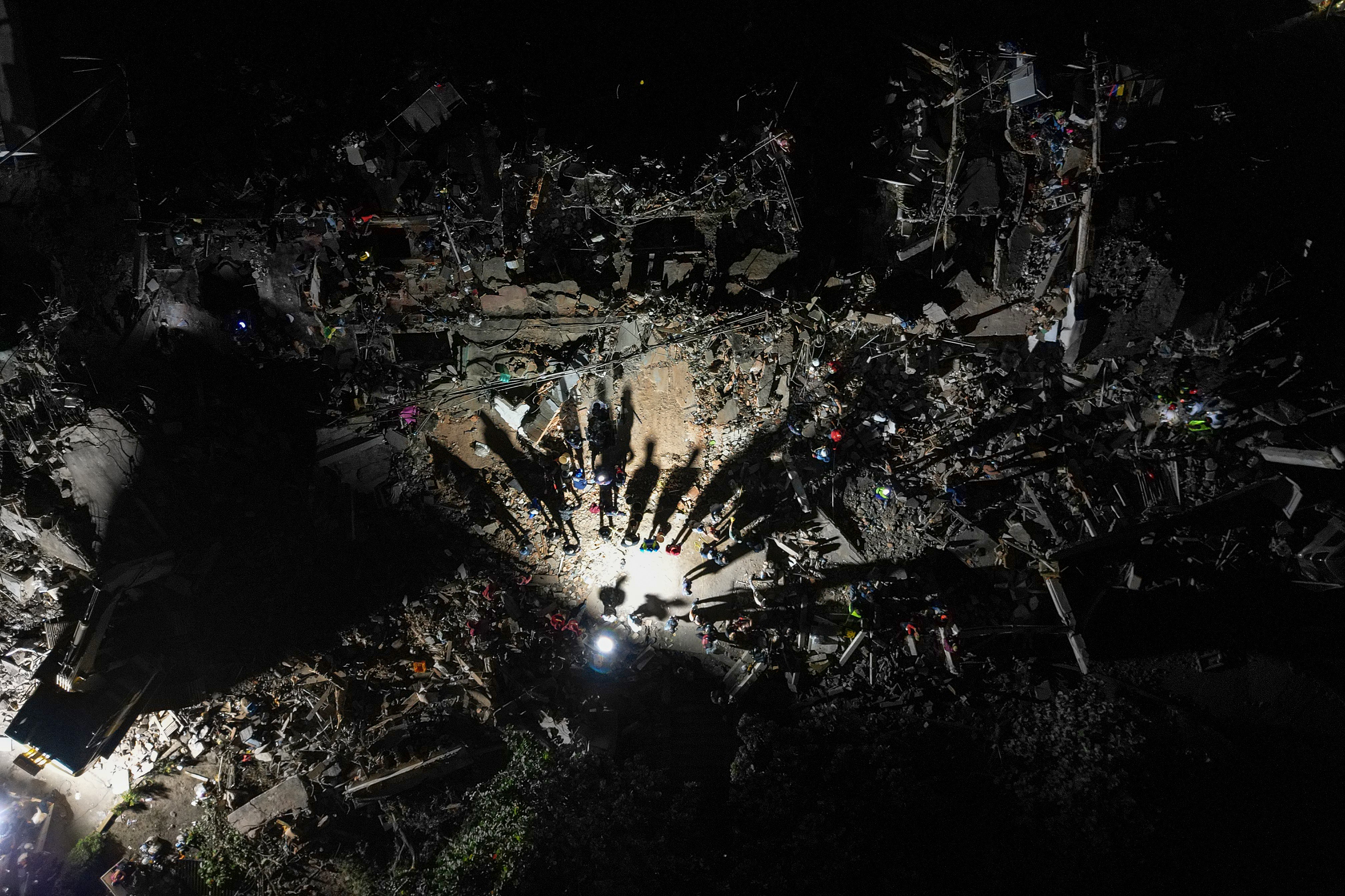 An aerial view of rescue workers amid destroyed buildings, seen at night, with long shadows cast by a work light.