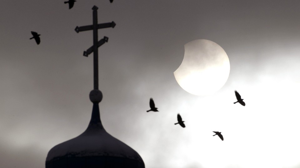Birds fly past a church spire in front of a partially eclipsed sun.