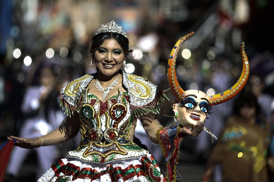 A person in costume dances in a parade while holding a horned mask.