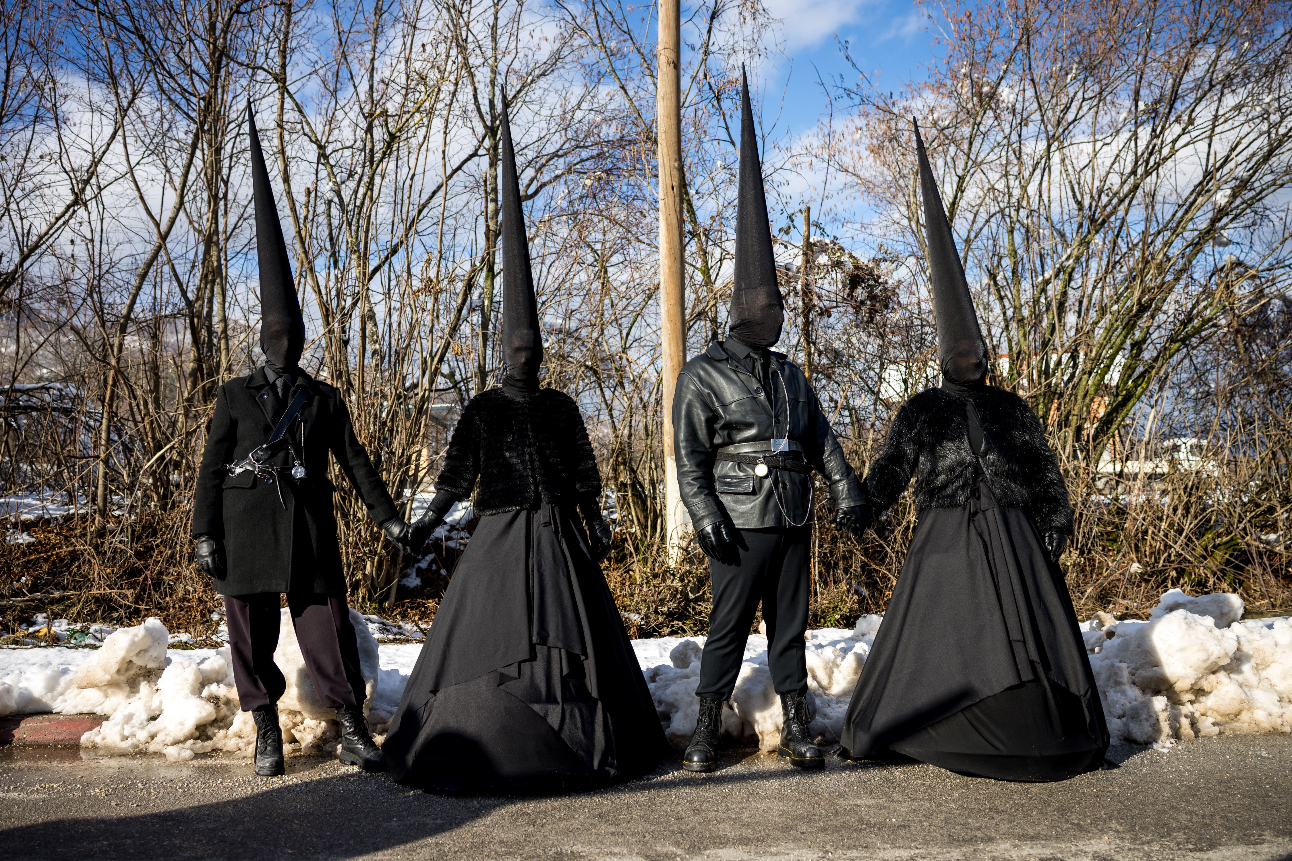 Two couples pose, all wearing costumes of black clothing, with tall conical black caps and black cloth over their heads.