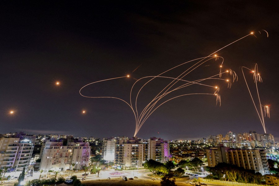 Rockets leave curving streaks of light in the night sky above buildings.