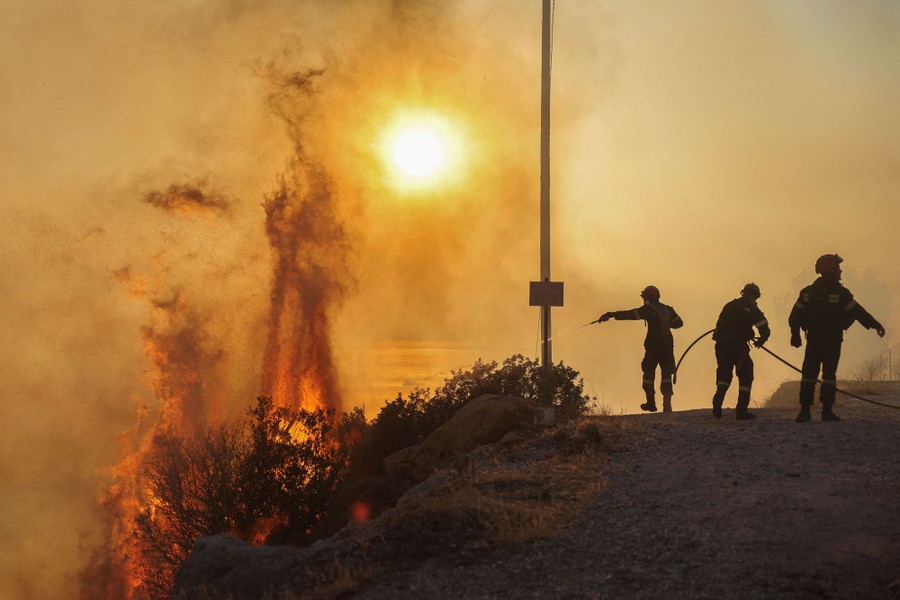 Several firefighters work with a hose on top of a hill, spraying water toward a brushfire below them.