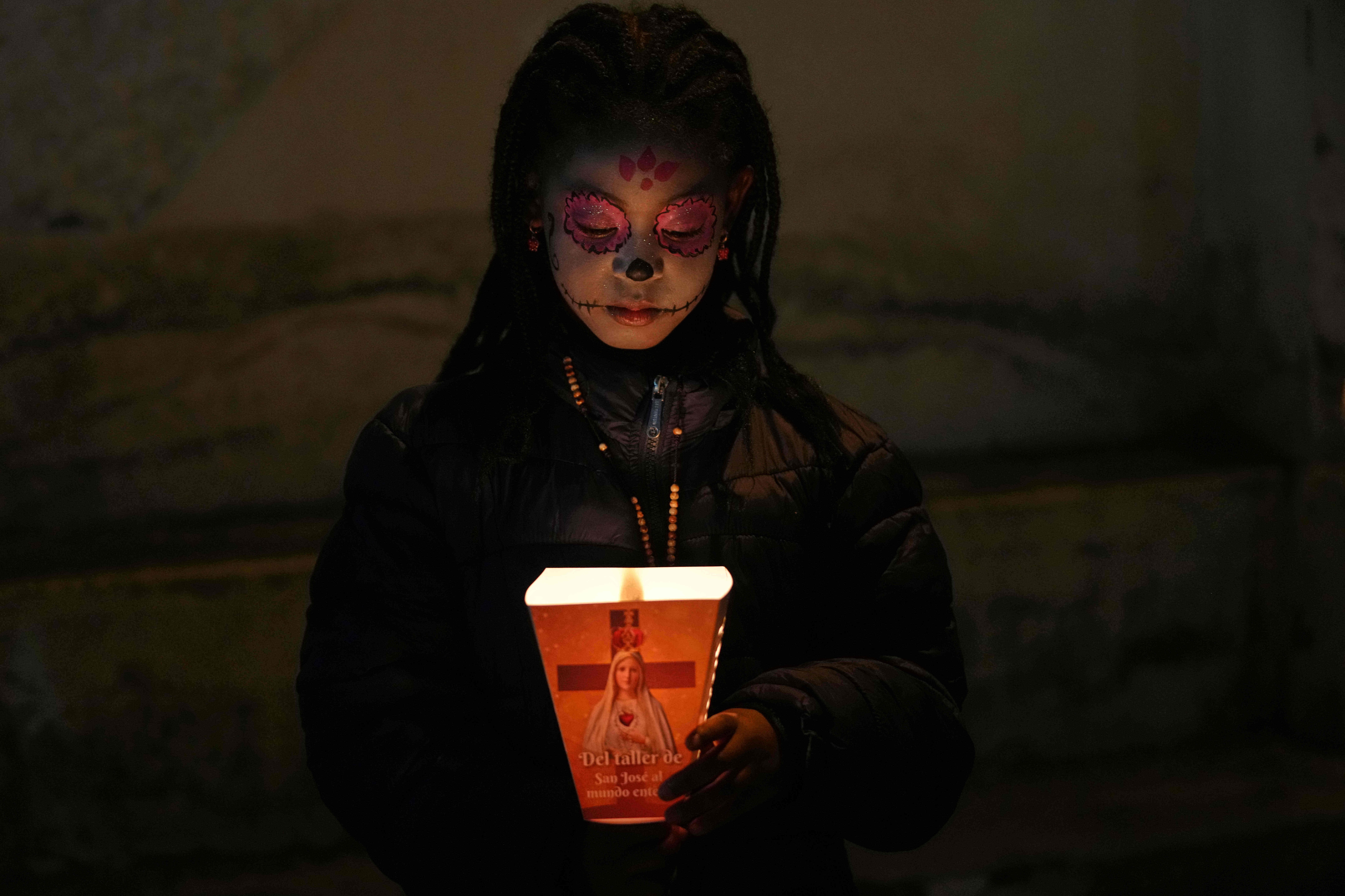 A person wearing traditional Catrina makeup looks down at a candle they are carrying during a Day of the Dead procession.