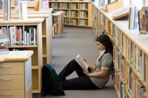 Asian American student sitting on the floor of a library reading a book, leaning back against the shelves