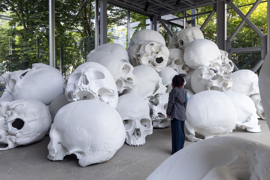 A person stands in front of an art installation made up of a couple dozen giant human skulls in a pile.