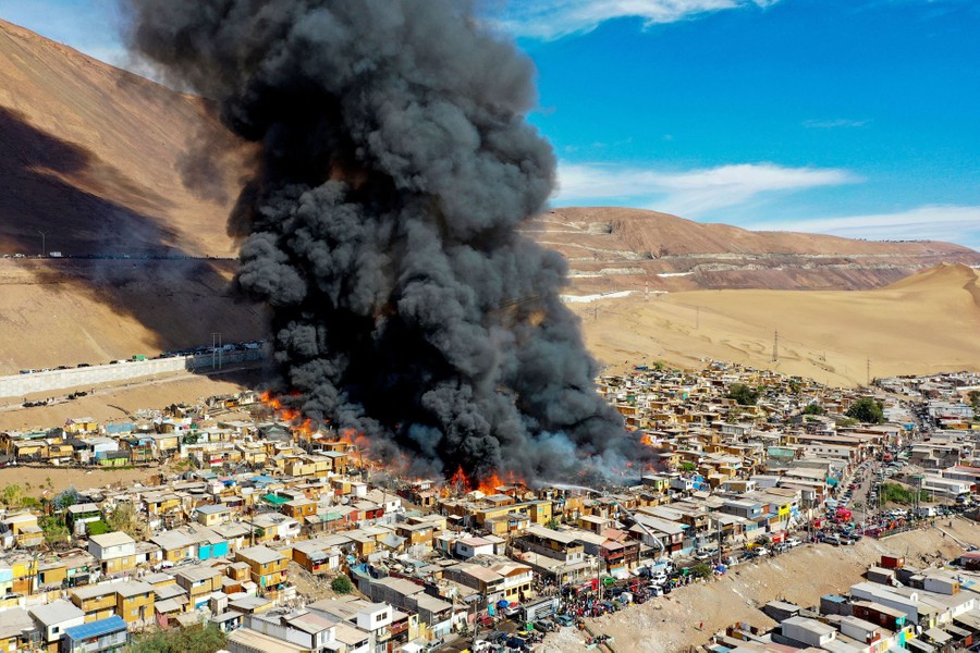 A large plume of smoke rises from burning buildings in a small residential area on a hillside.